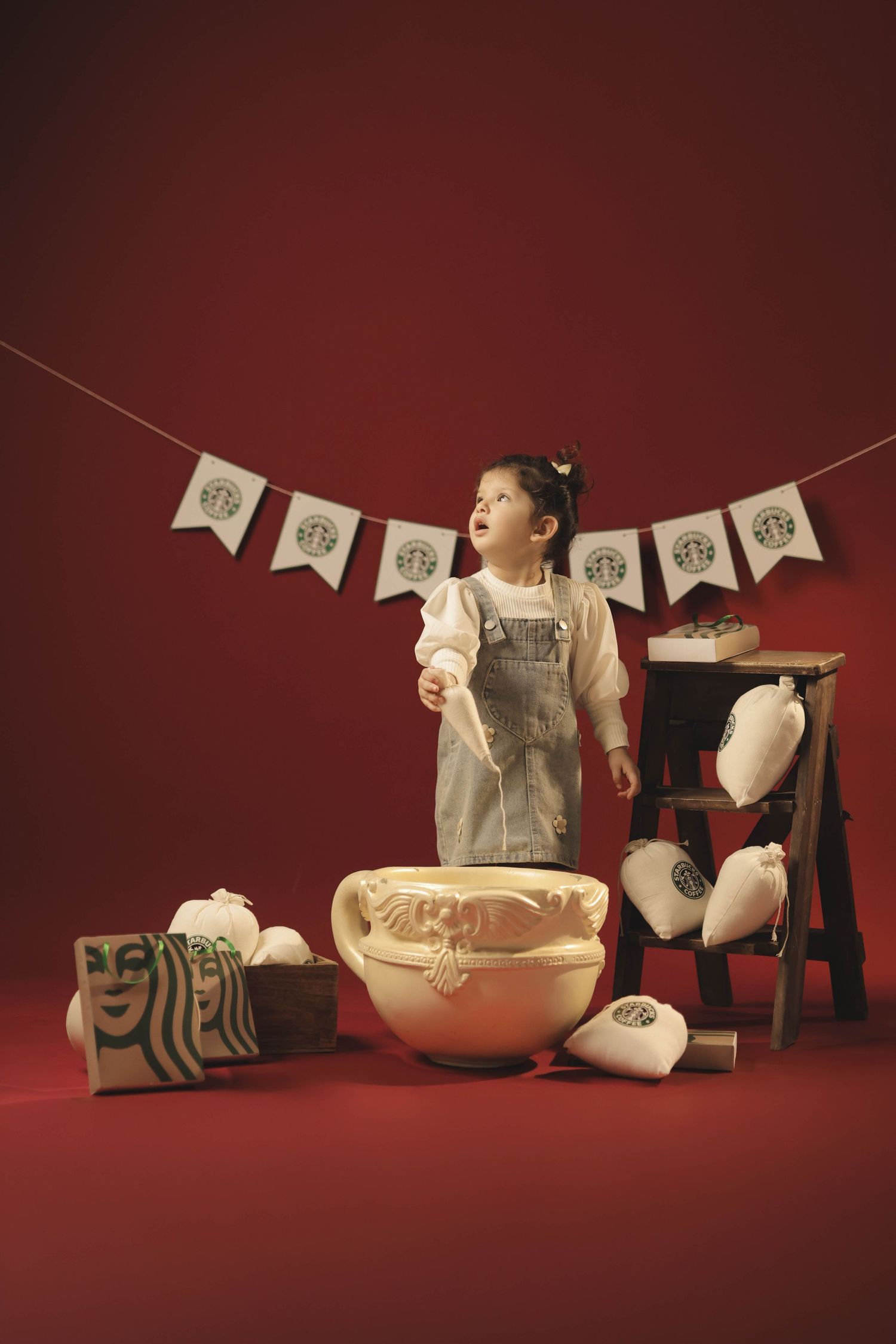 A toddler girl stands among Starbucks-themed decorations, plush toys, and a large bowl on a red background.