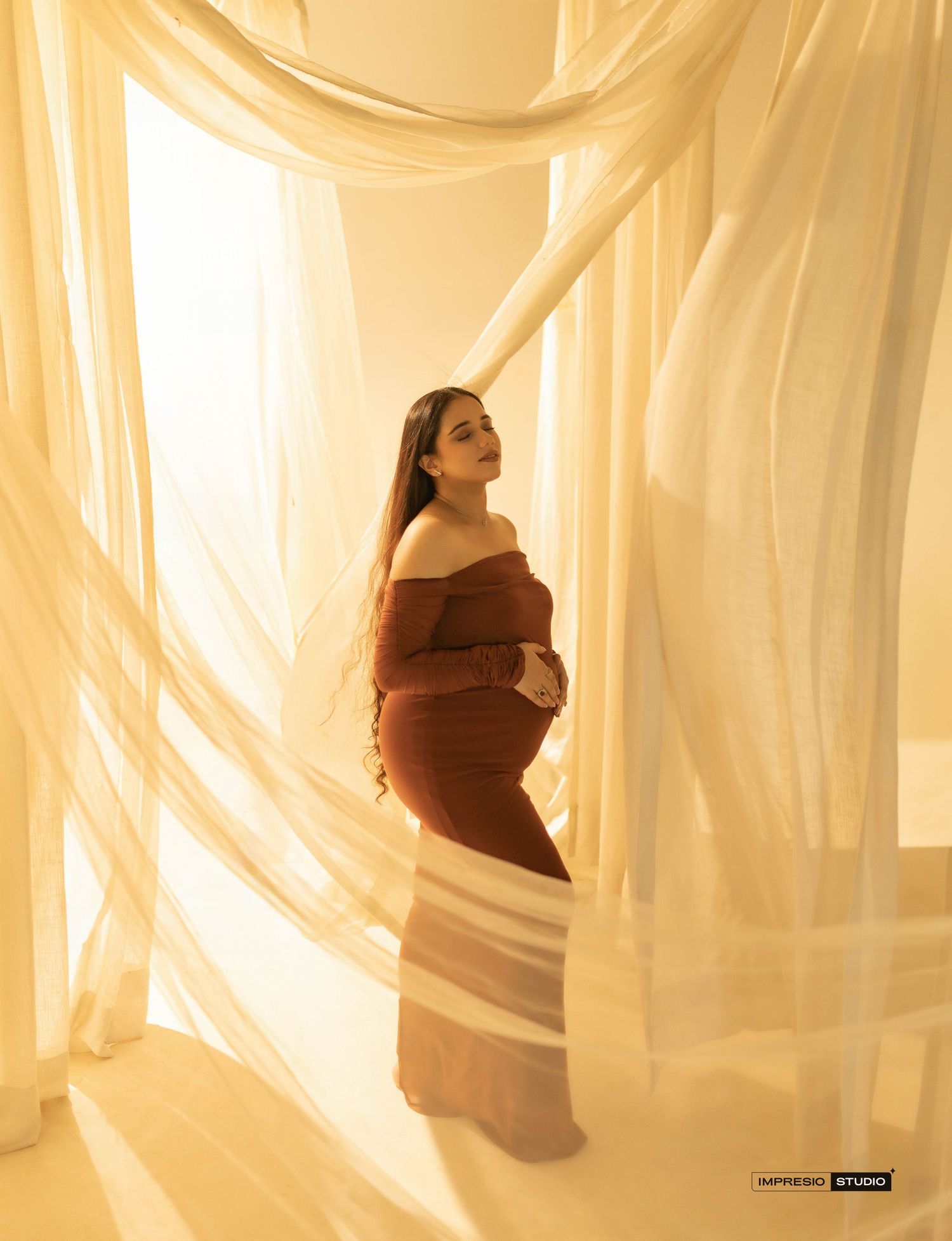 Pregnant woman in rust off-shoulder gown poses gracefully among flowing sheer curtains in warm golden light.