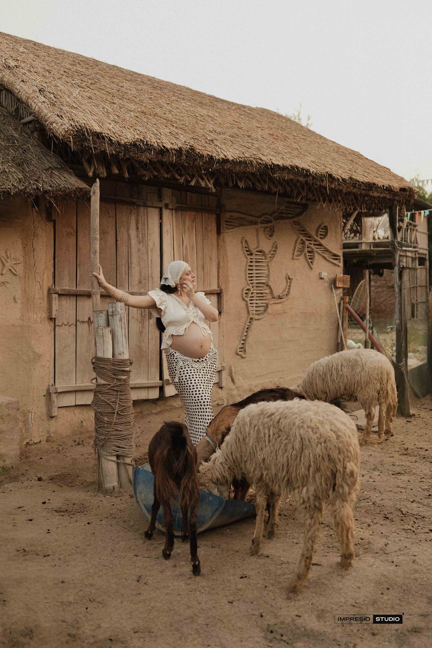 Pregnant woman in polka dot skirt feeding sheep and goats near rustic mud hut with thatched roof on a farm.