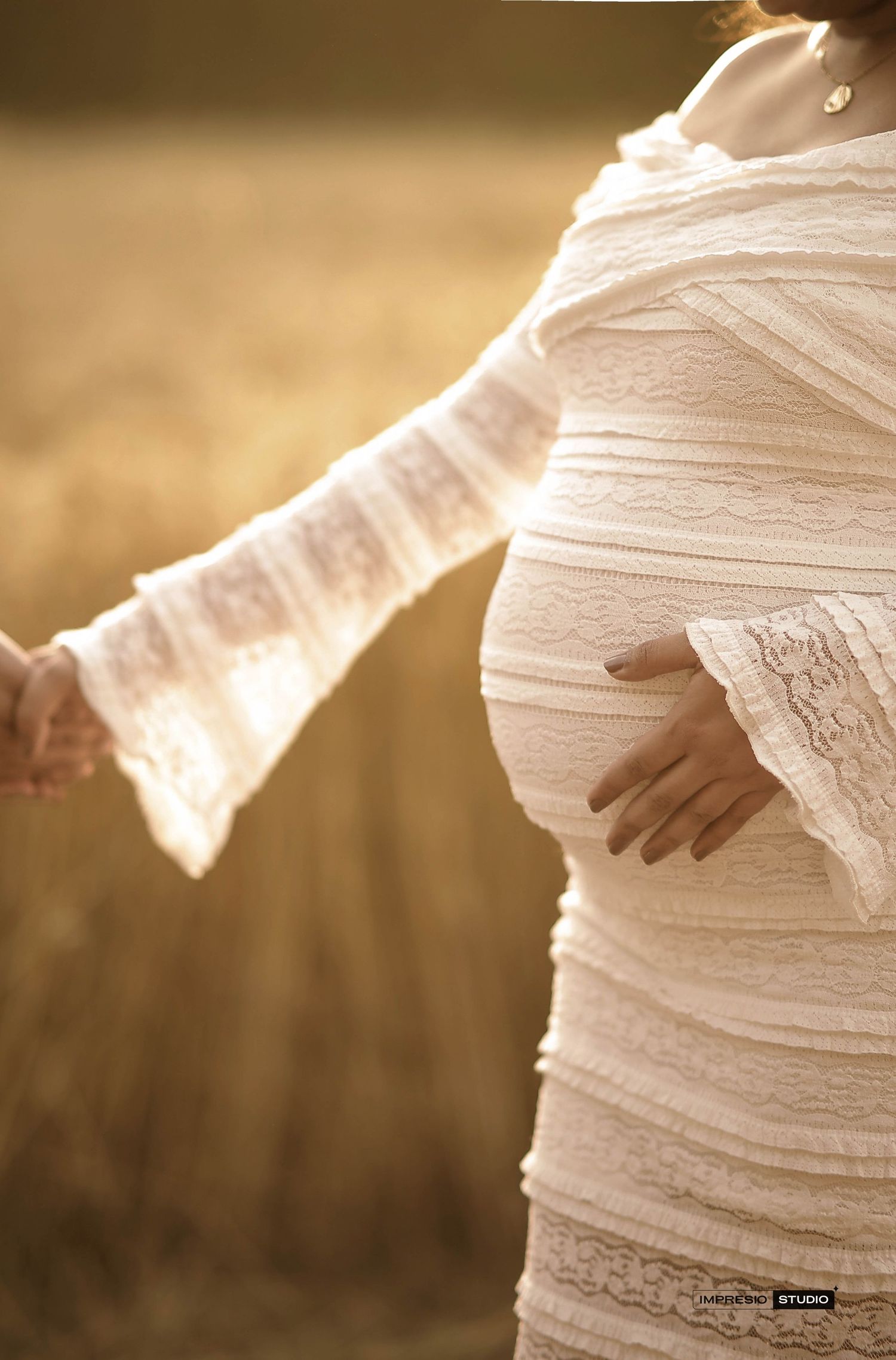Pregnant woman in white lace dress holding hands in golden field, cradling baby bump during maternity photoshoot.