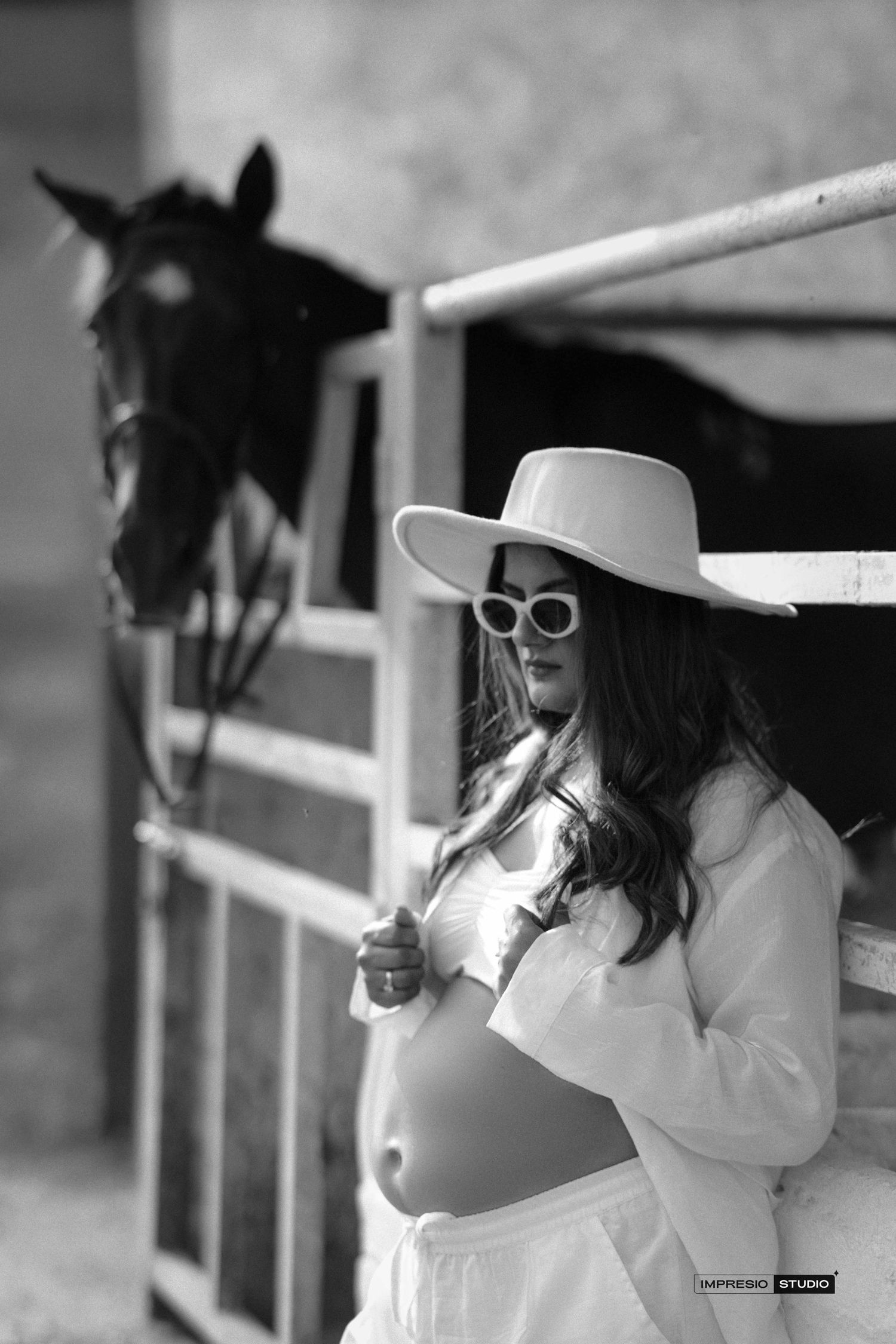 Black and white maternity photo of pregnant woman in white hat and sunglasses posing near horse stable.