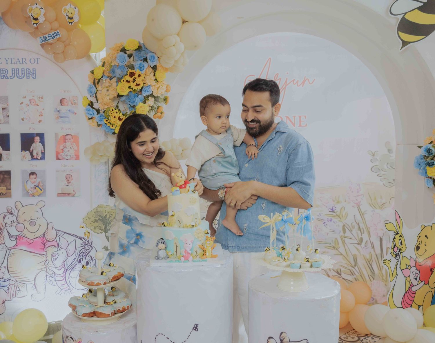 Family celebrating baby's first birthday with Winnie the Pooh themed cake and balloon decorations.