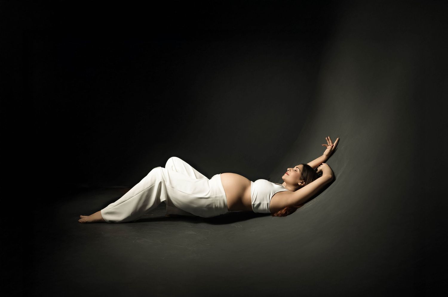 Pregnant woman in white dress lying gracefully on dark floor with arm extended, artistic maternity photography.