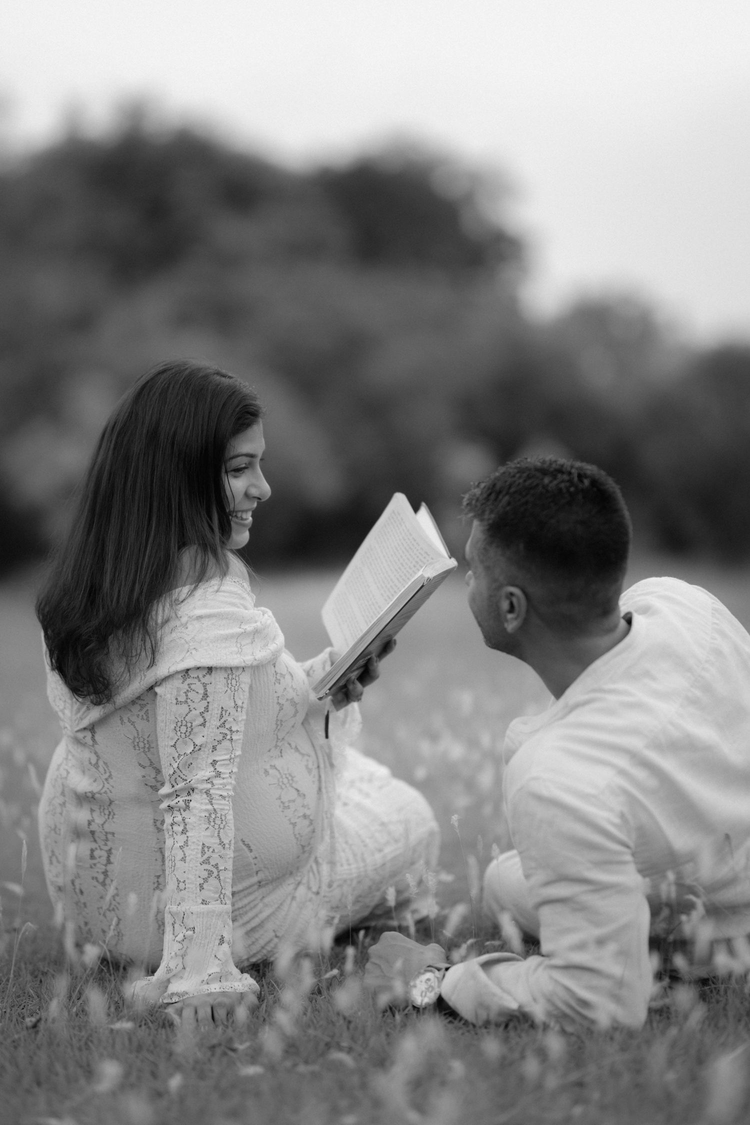 Black and white photo of a smiling couple sitting in a field, sharing a book together on a romantic outdoor date.