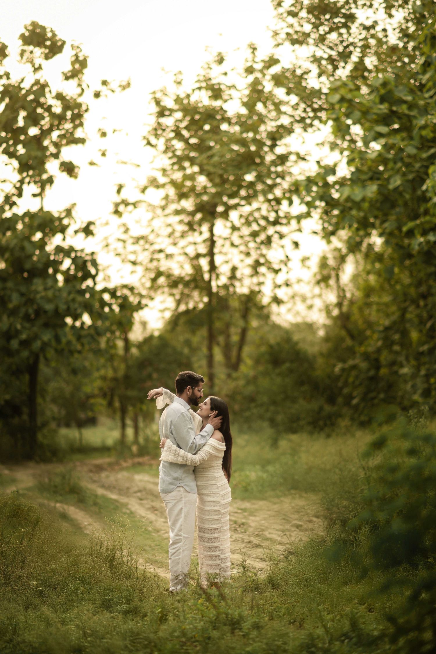 Romantic couple embracing on a rustic dirt path surrounded by lush green trees in golden sunset light.