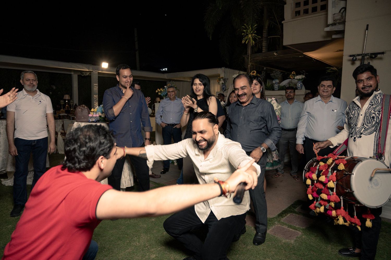 Men dancing energetically at an outdoor evening celebration with live dhol drummer and guests watching.