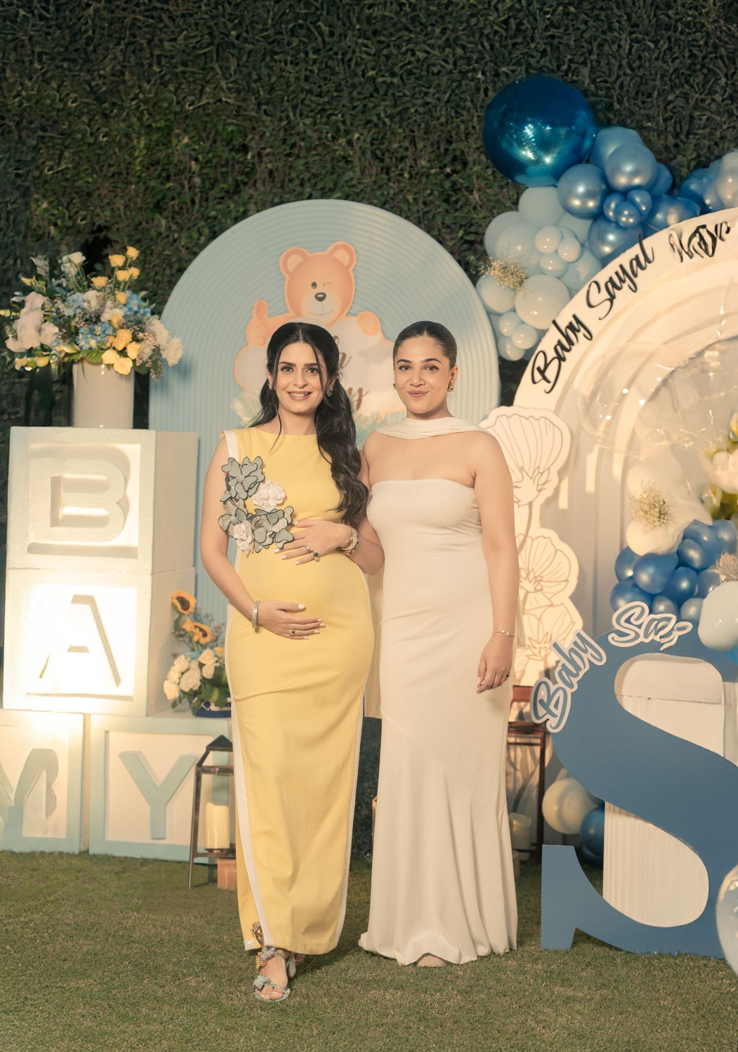 Two pregnant women in elegant dresses pose at a baby shower decorated with blue balloons and teddy bear themed backdrop.