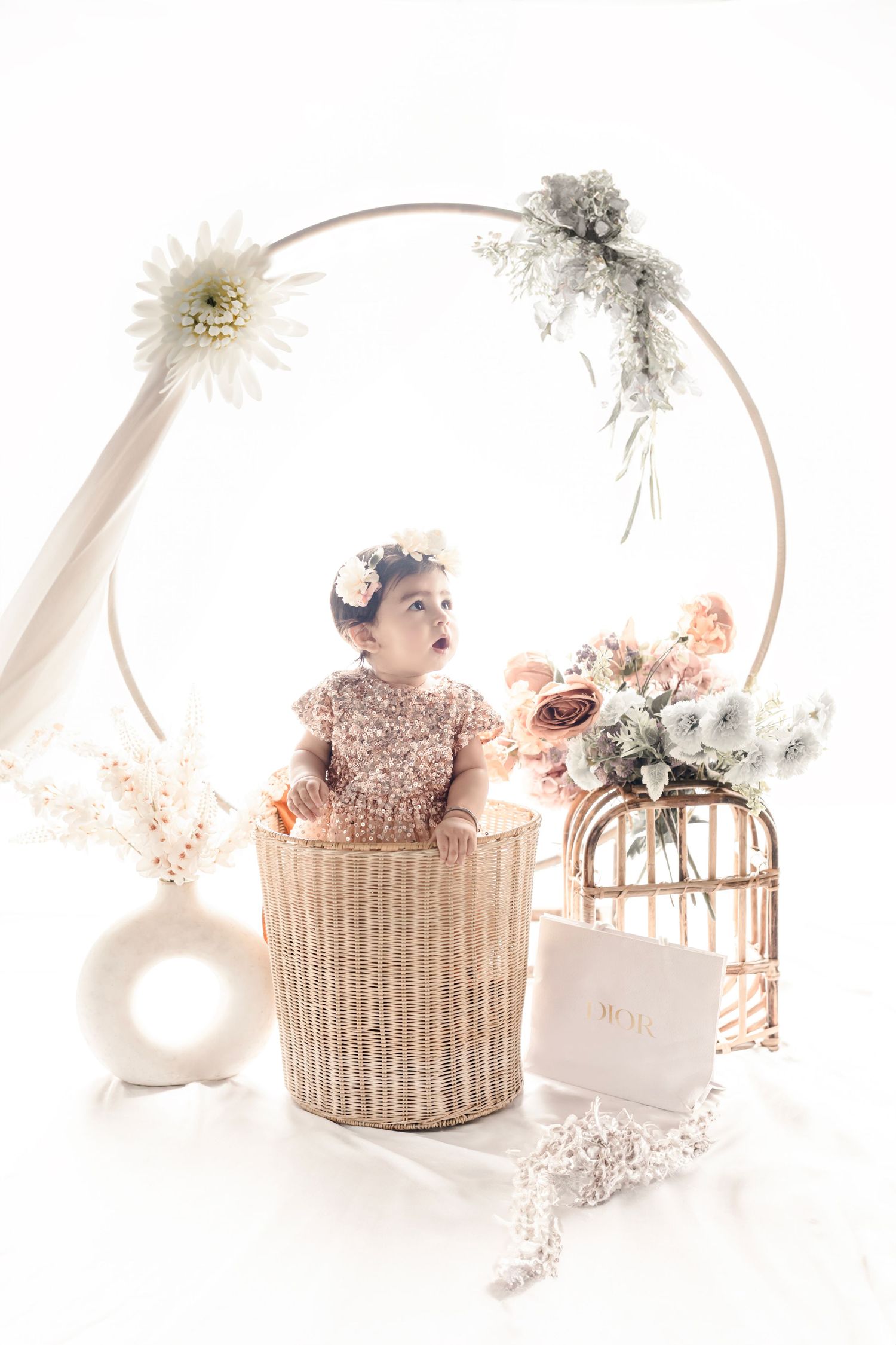 Baby girl in sparkly pink outfit sits in wicker basket surrounded by flowers and floral hoop backdrop.