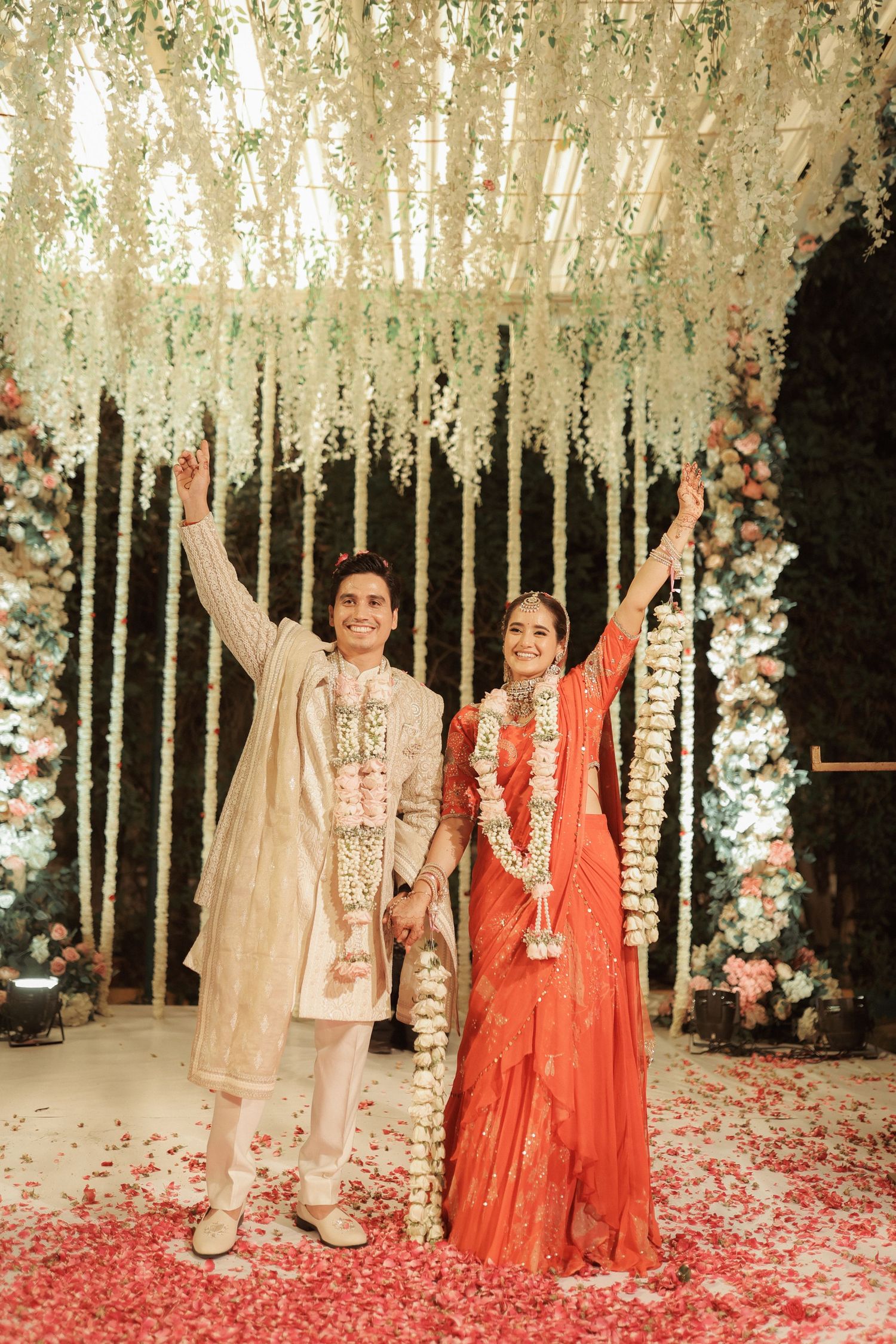 Indian couple in traditional attire celebrate wedding under floral canopy with raised hands.