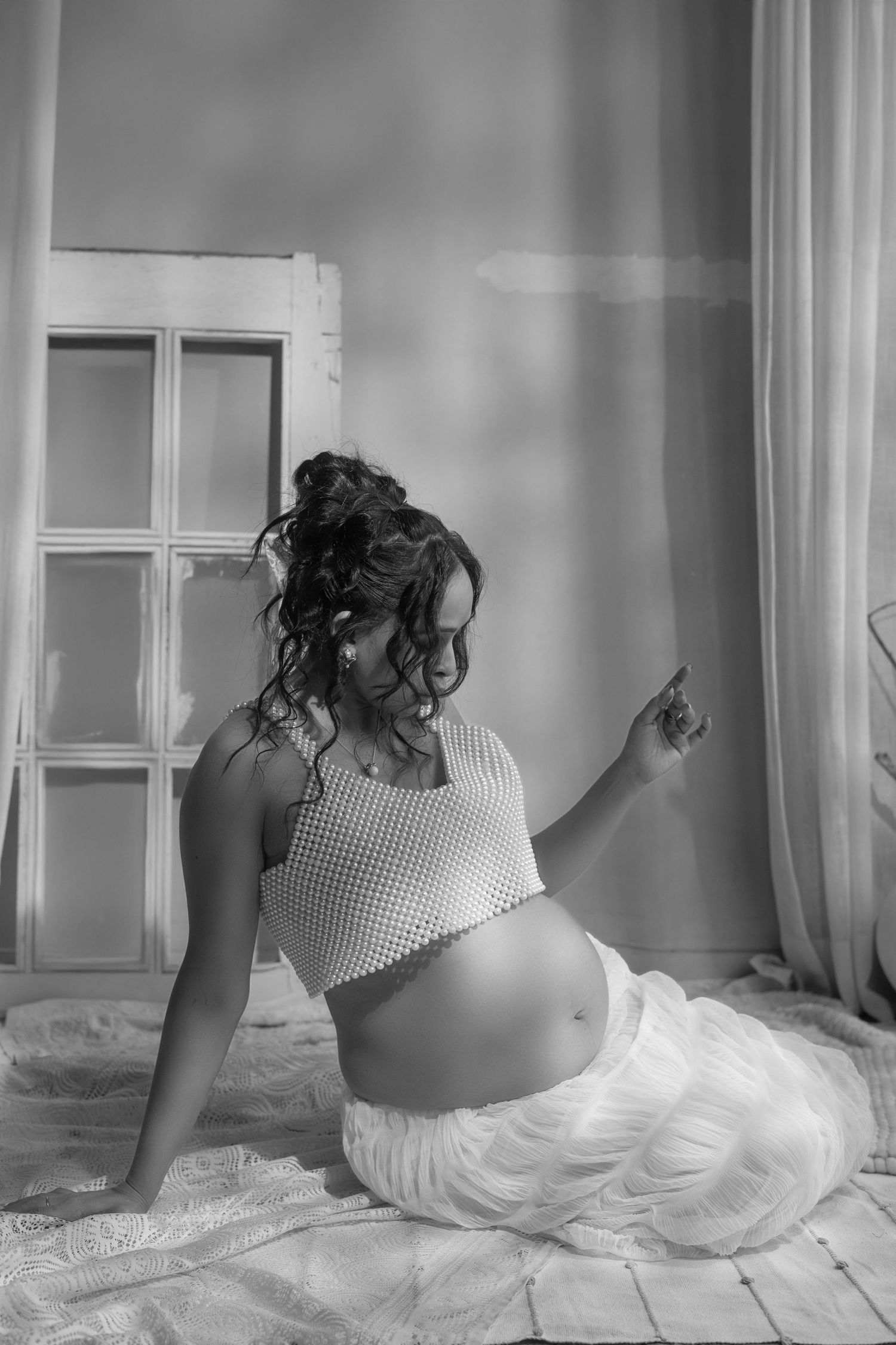 Black and white maternity photo of pregnant woman in crop top and white skirt sitting gracefully on floor near window.