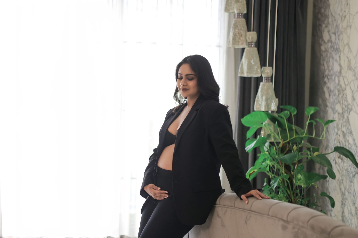 Casual maternity photo with black blazer near indoor plants and window.