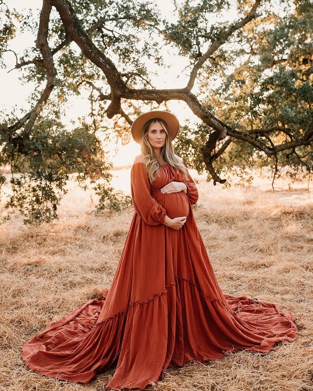 Sunset maternity portrait with flowing rust-colored dress and wide brim hat in outdoor field setting.