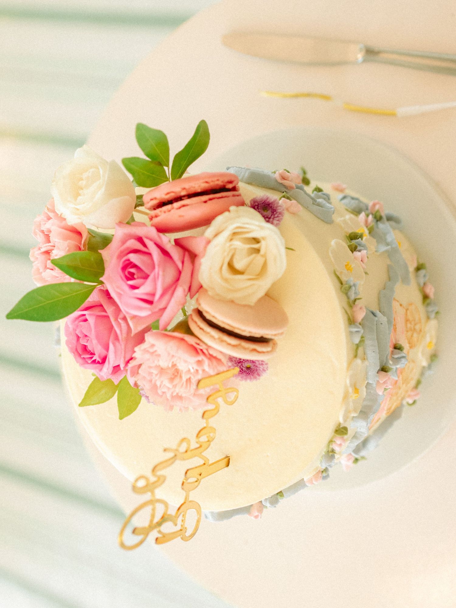 Elegant white wedding cake topped with pink roses, white ranunculus, and macarons, decorated with gold script topper.