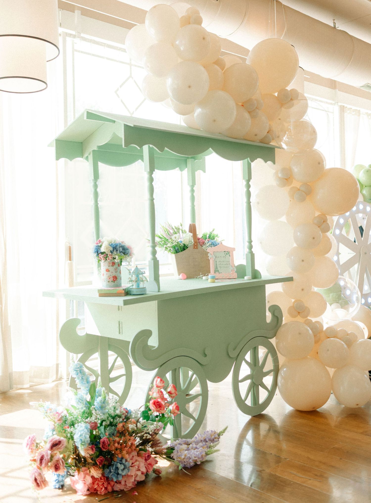 Mint green decorative candy cart adorned with white balloon arch and colorful floral arrangements at an elegant party.