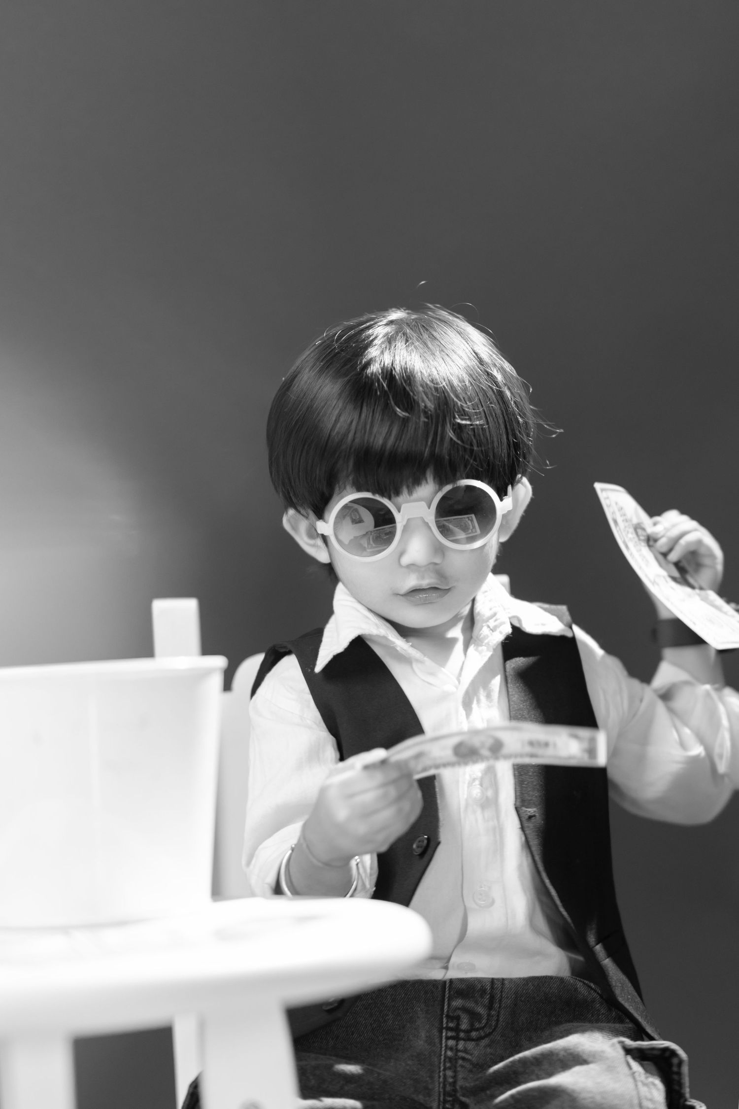 Black and white photo of a young boy wearing round glasses, holding paper money while sitting at a white table.