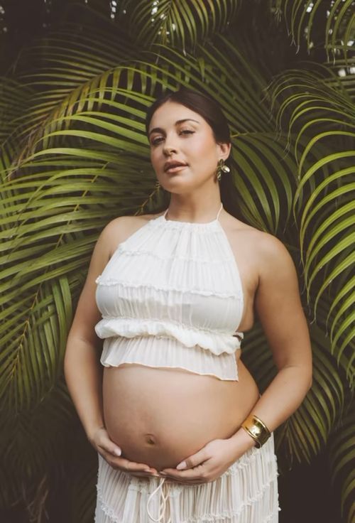 A woman in a white halter top poses against tropical palm fronds during a maternity photoshoot.