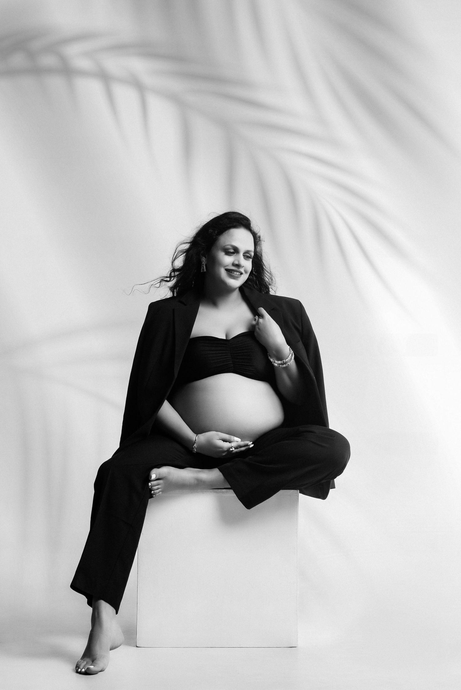 A black and white maternity portrait with palm shadow effects and minimalist studio setting.