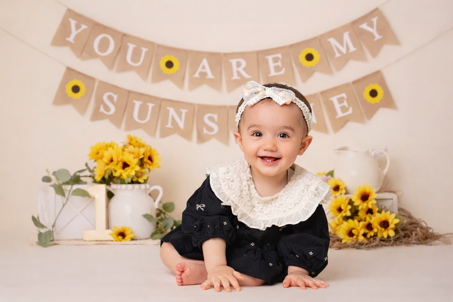 Baby in black dress and white headband poses with yellow sunflowers and banner decorations at photo session.