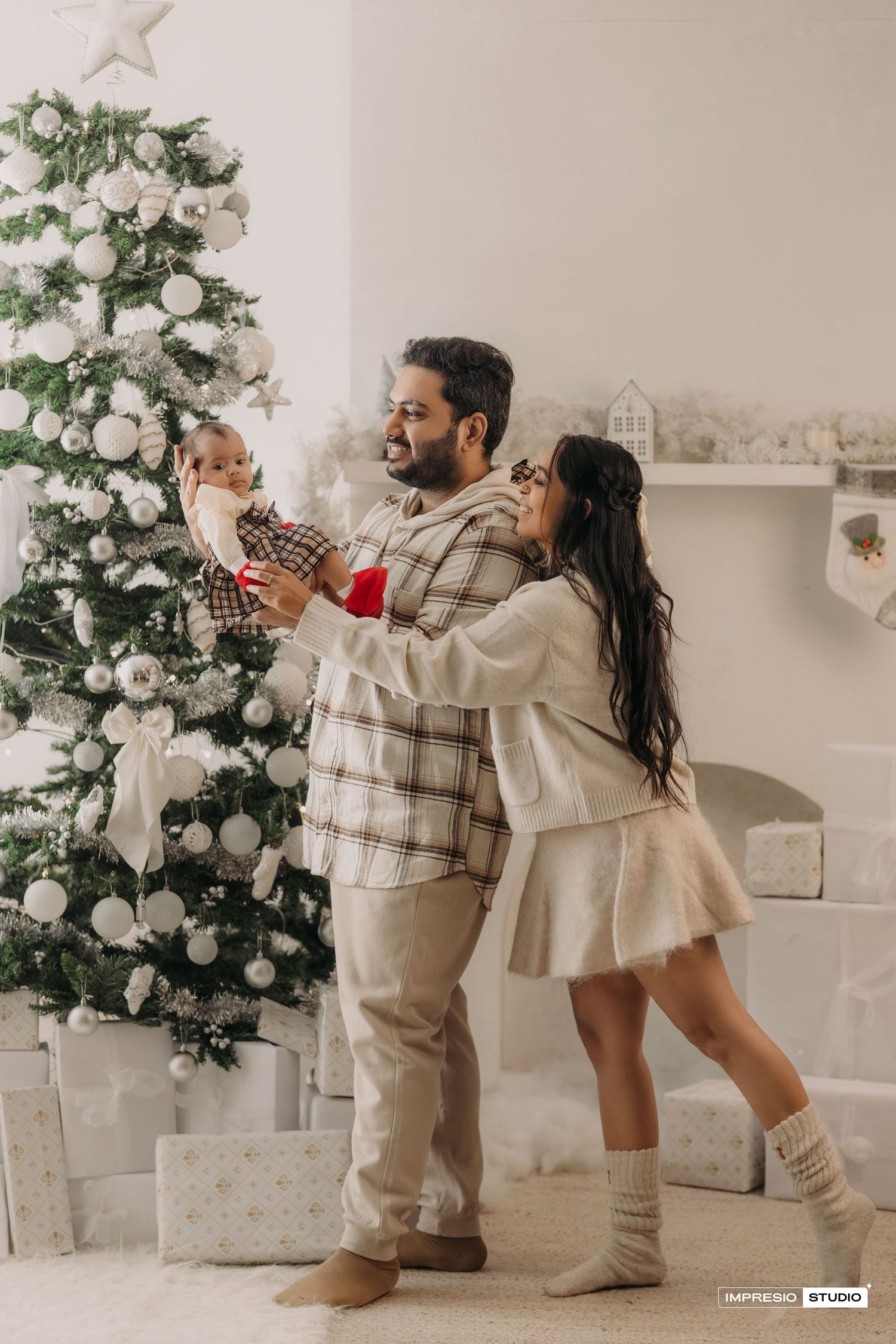 A couple in cozy winter outfits decorates a white Christmas tree together in a bright modern living room.