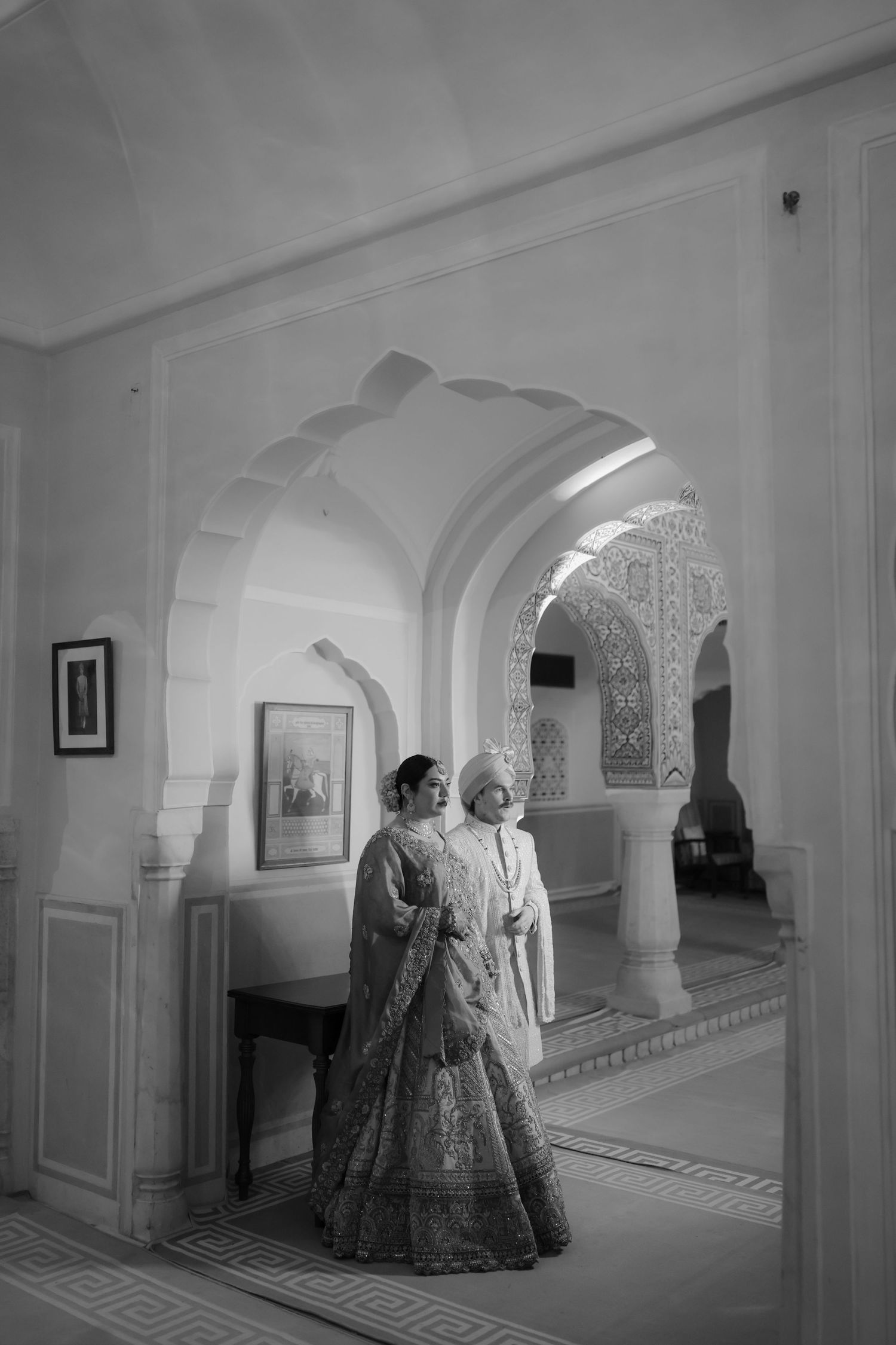 Vintage black and white photograph of people walking through an ornate corridor with arched doorways and detailed architecture.