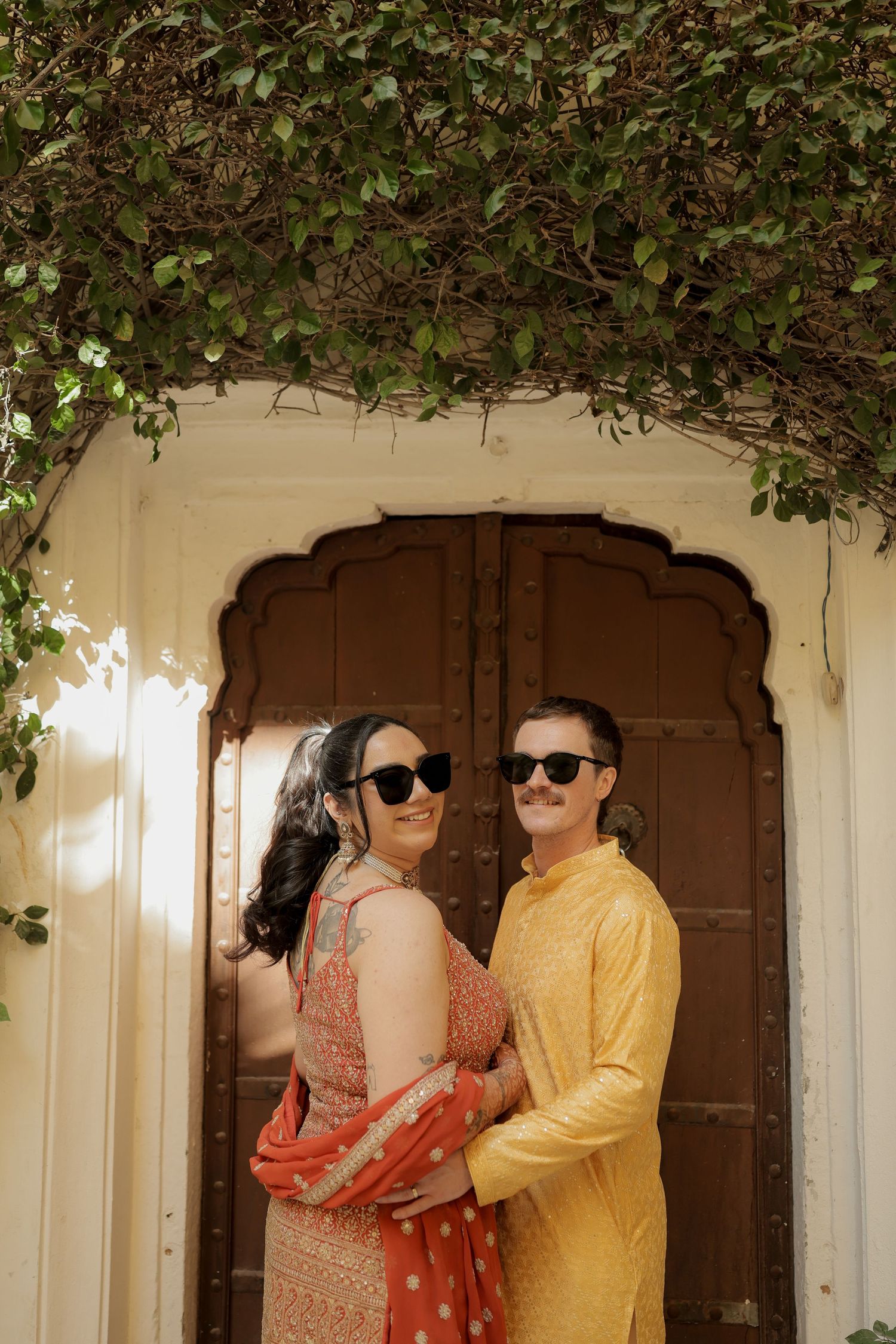 Couple in sunglasses posing against white archways decorated with green vines.