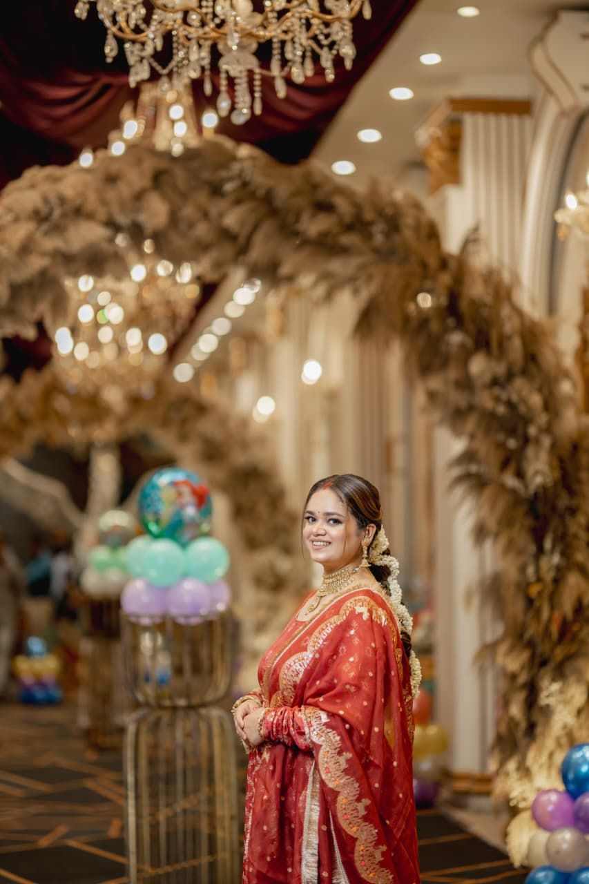 A bride in a red and gold embroidered saree smiles at a decorated wedding venue with chandeliers and floral arches.
