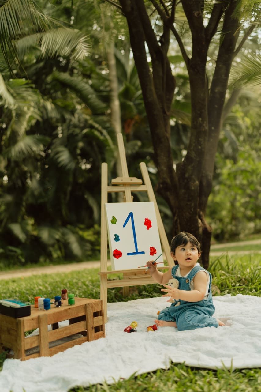 Toddler girl in blue dress sits on white blanket outdoors beside a painted canvas showing number 1 for first birthday.