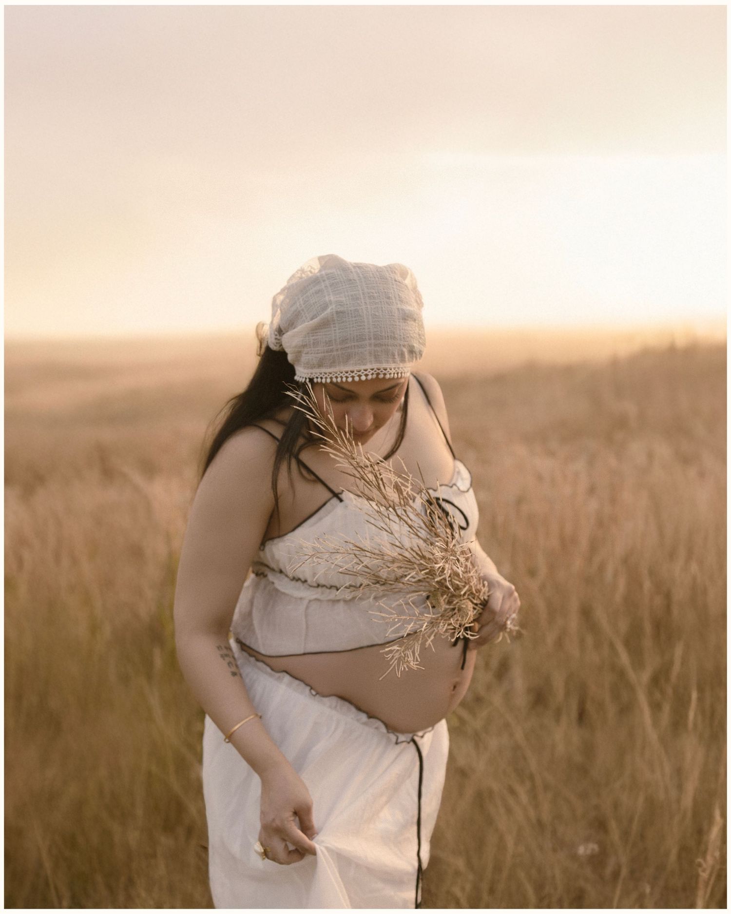 Pregnant woman in white outfit holding dried wheat in golden field at sunset.