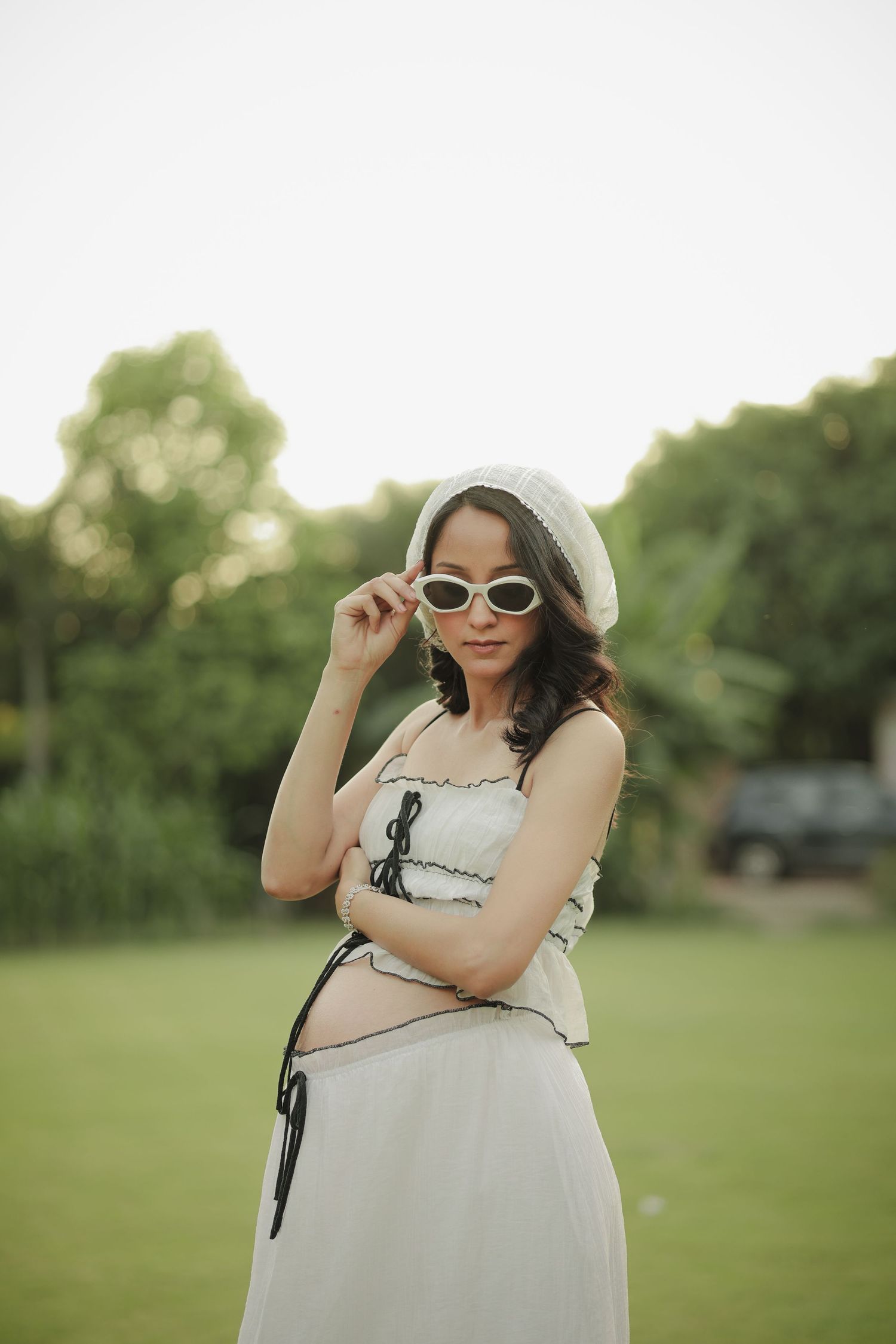 Pregnant woman in white beret and sunglasses posing in a green park wearing a stylish white crop top and skirt.