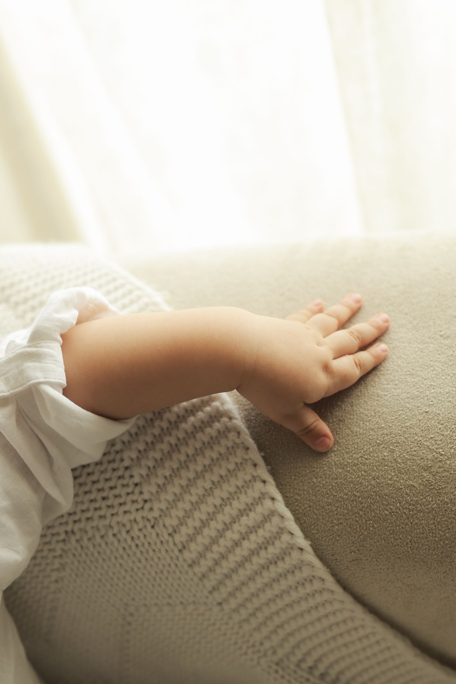Close-up of a baby's small hand resting on a beige sofa cushion, wearing a white long-sleeve outfit.