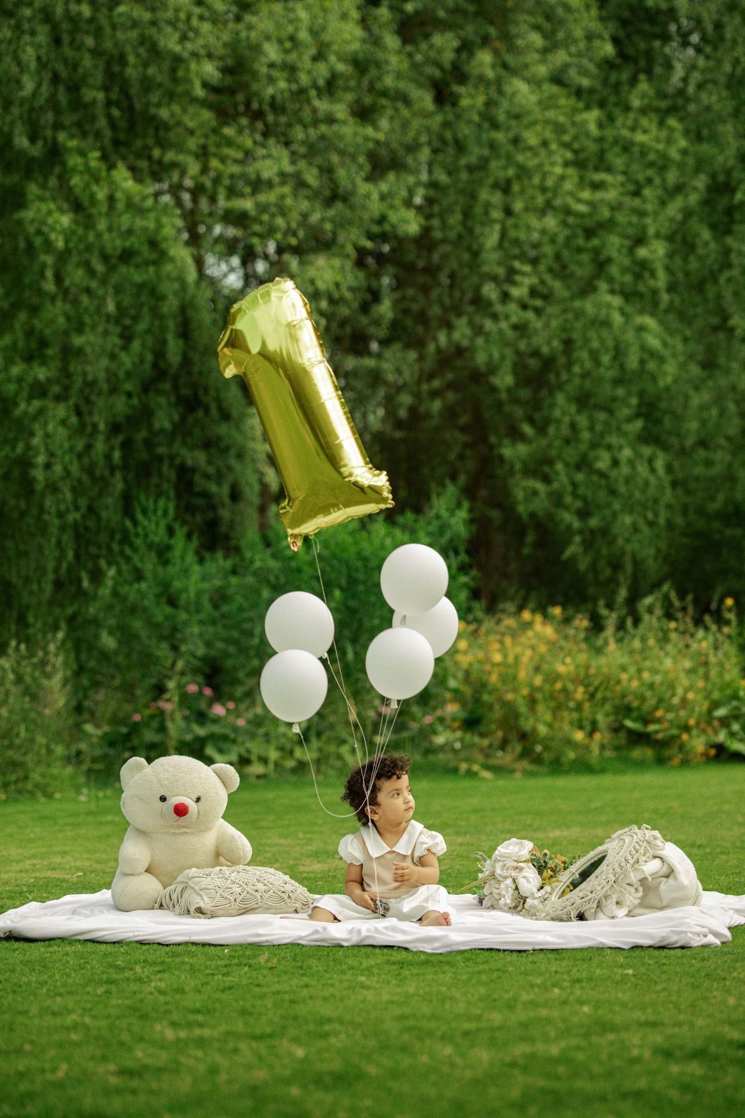 Baby's first birthday celebration on a blanket in a park with a gold number 1 balloon, white balloons, and a teddy bear.