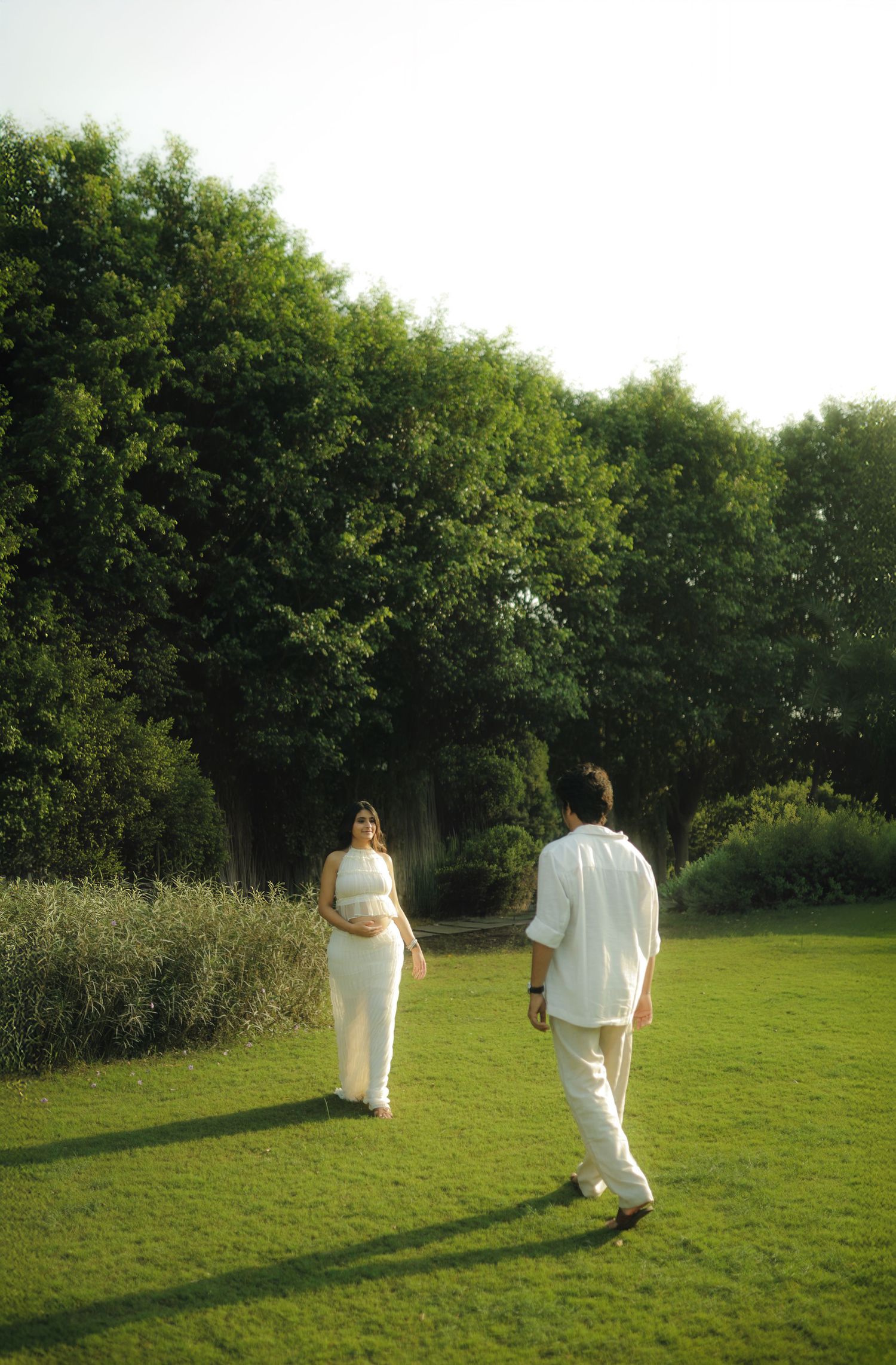 Couple in white outfits walking on green lawn surrounded by lush trees in a garden setting.