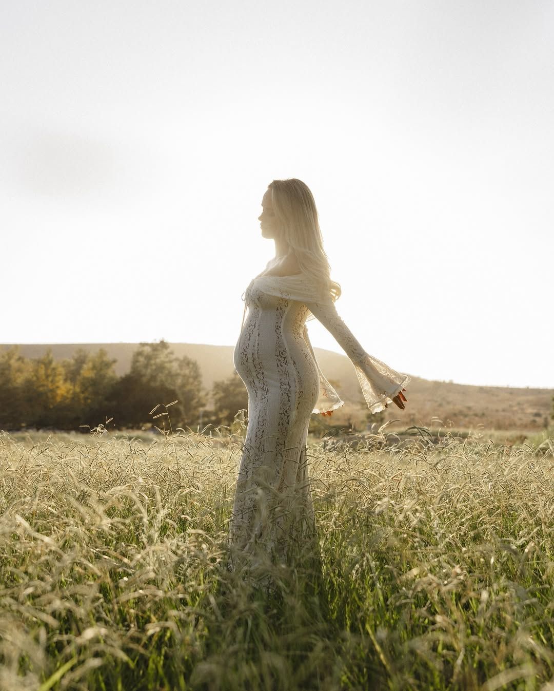 Ethereal silhouette of expectant mother in lace dress amid golden sunlit field.