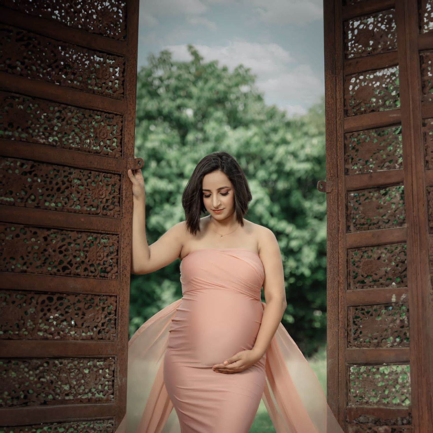 Maternity portrait in pink dress framed by ornate wooden doors in garden setting.