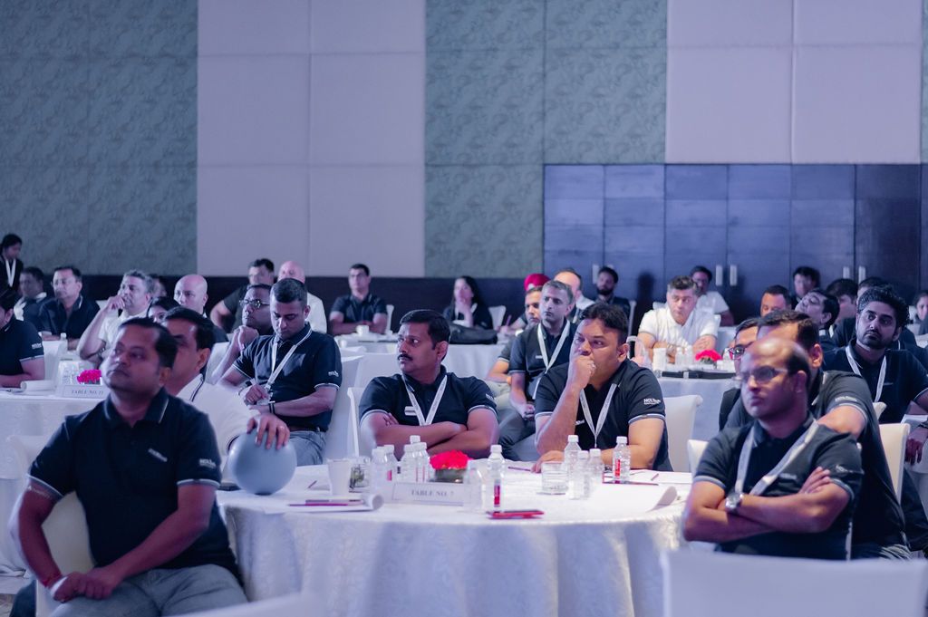 Conference attendees sit at round tables in a large meeting room during a business event.