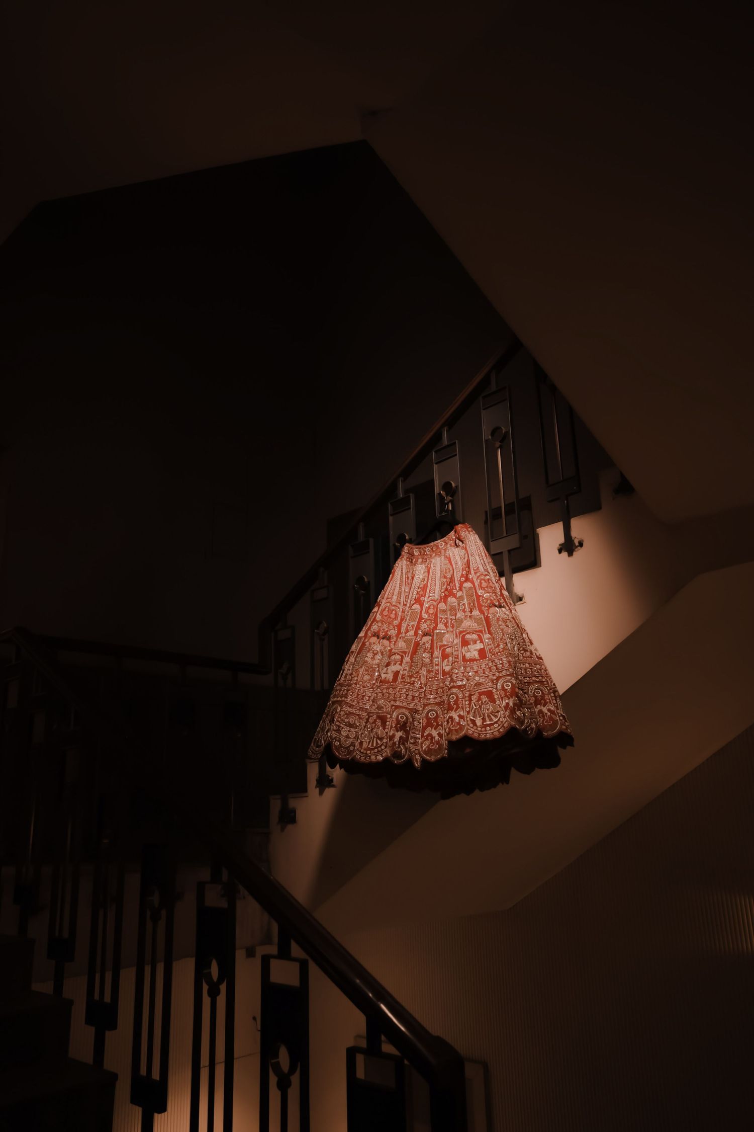 Ornate red and white embroidered lehenga skirt draped over dark staircase railing in dim warm lighting.