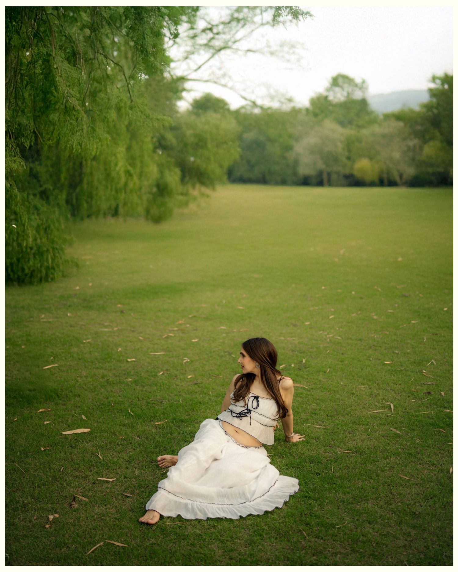 Woman in white dress sitting on lush green grass under willow tree in scenic outdoor setting.
