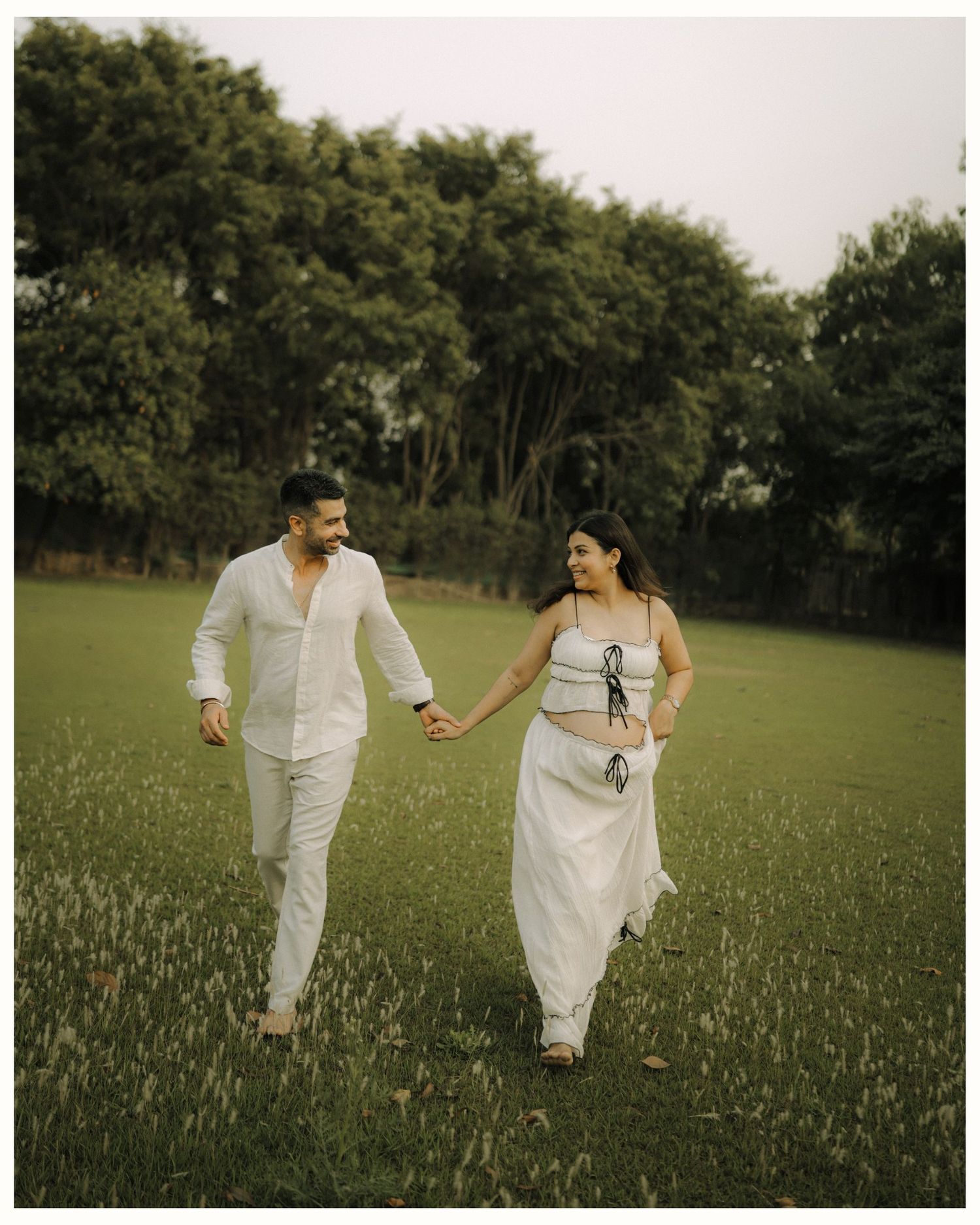 Couple in white outfits holding hands while walking through a lush green field with trees in the background.