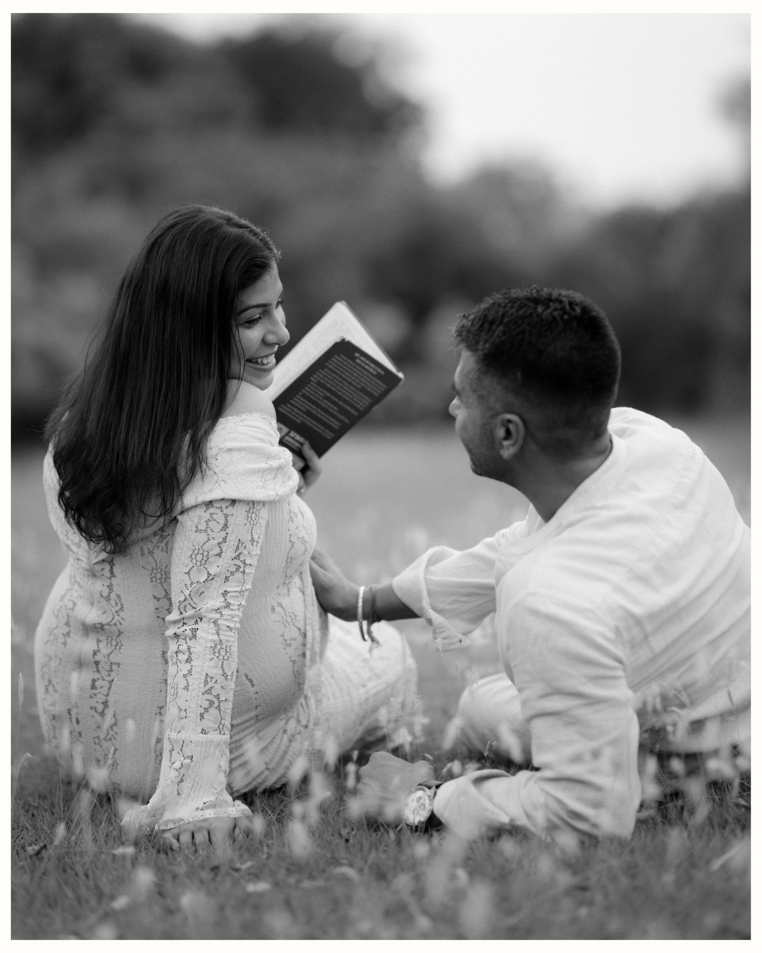 Black and white photo of a smiling couple sitting in grass, woman holding a book while man looks at her adoringly.