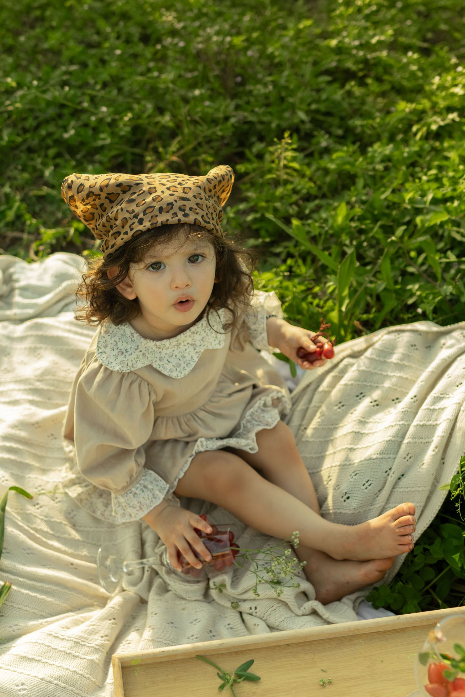 A toddler girl in a beige lace dress and leopard print hat sits barefoot on a blanket outdoors.