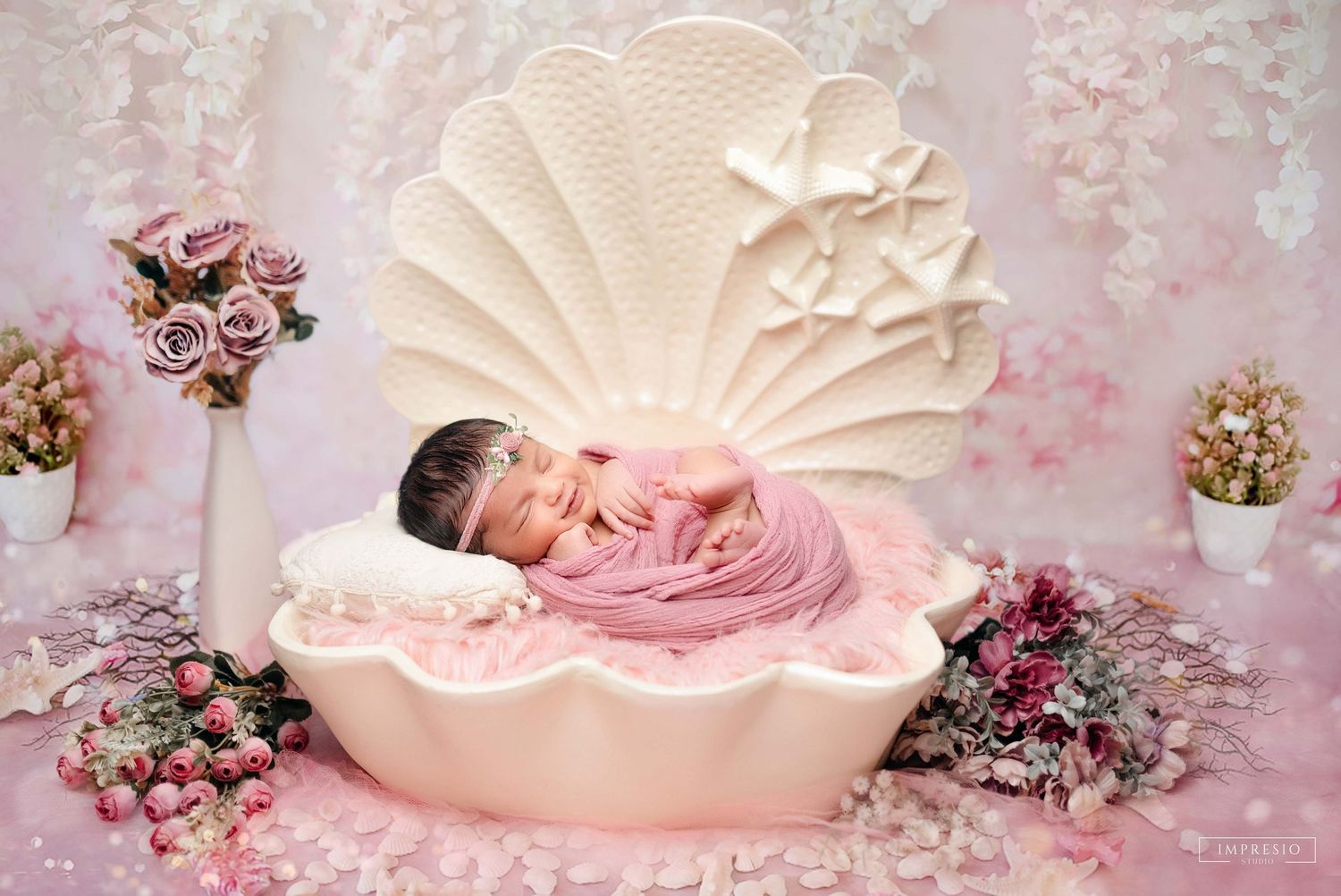 Newborn baby sleeping peacefully in a pink seashell prop surrounded by flowers in a dreamy pink studio setup.