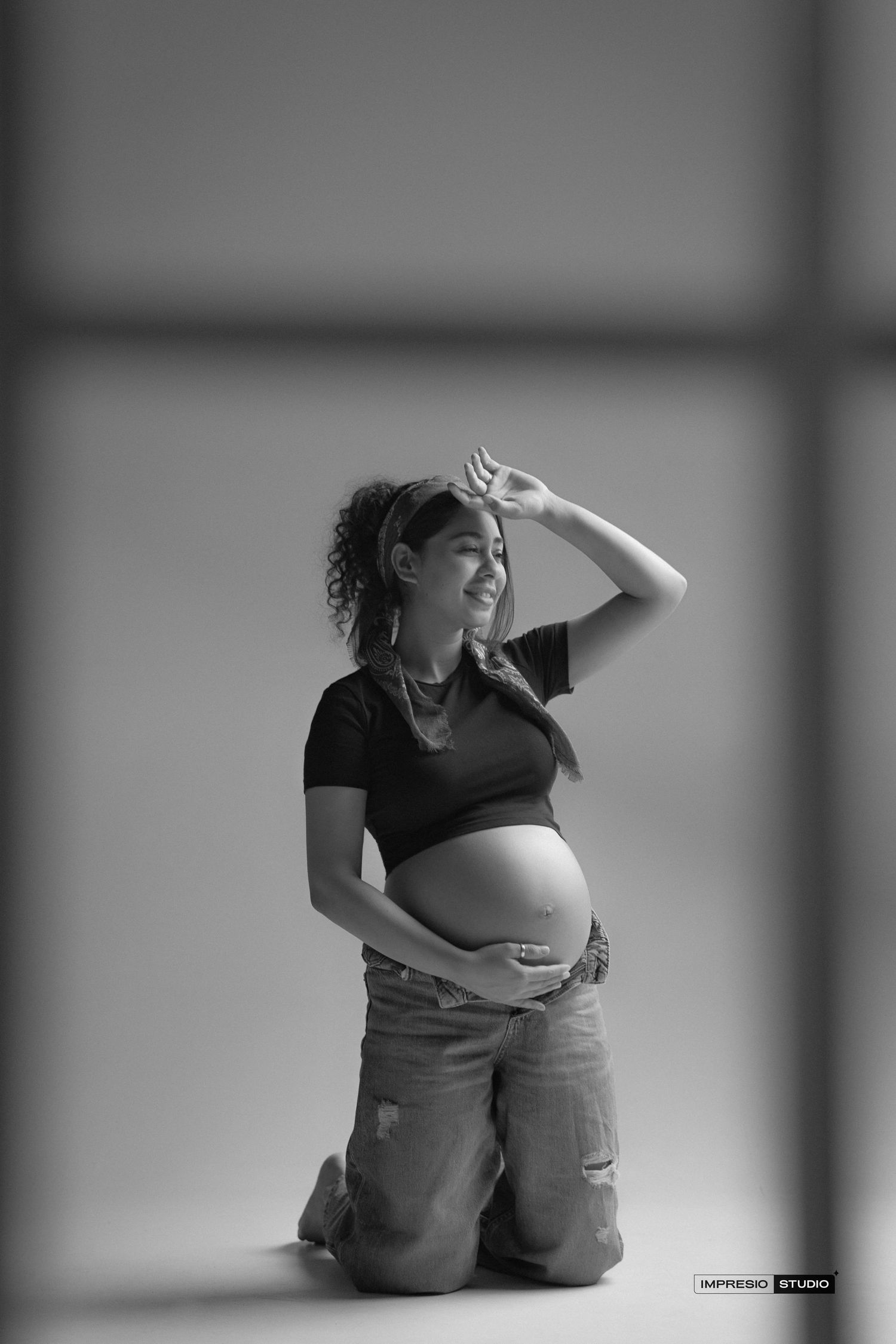 Black and white photo of a smiling pregnant woman kneeling, cradling her baby bump in a black top and ripped jeans.