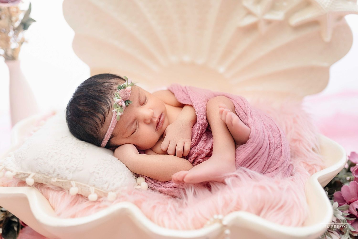 Newborn baby sleeping peacefully in a pink shell prop wearing a floral headband and pink wrap for a dreamy photoshoot.