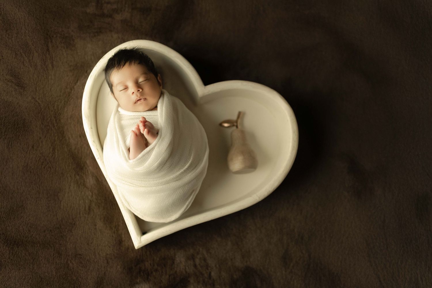 Newborn baby wrapped in white swaddle sleeping peacefully inside a heart-shaped white bowl on brown fur.