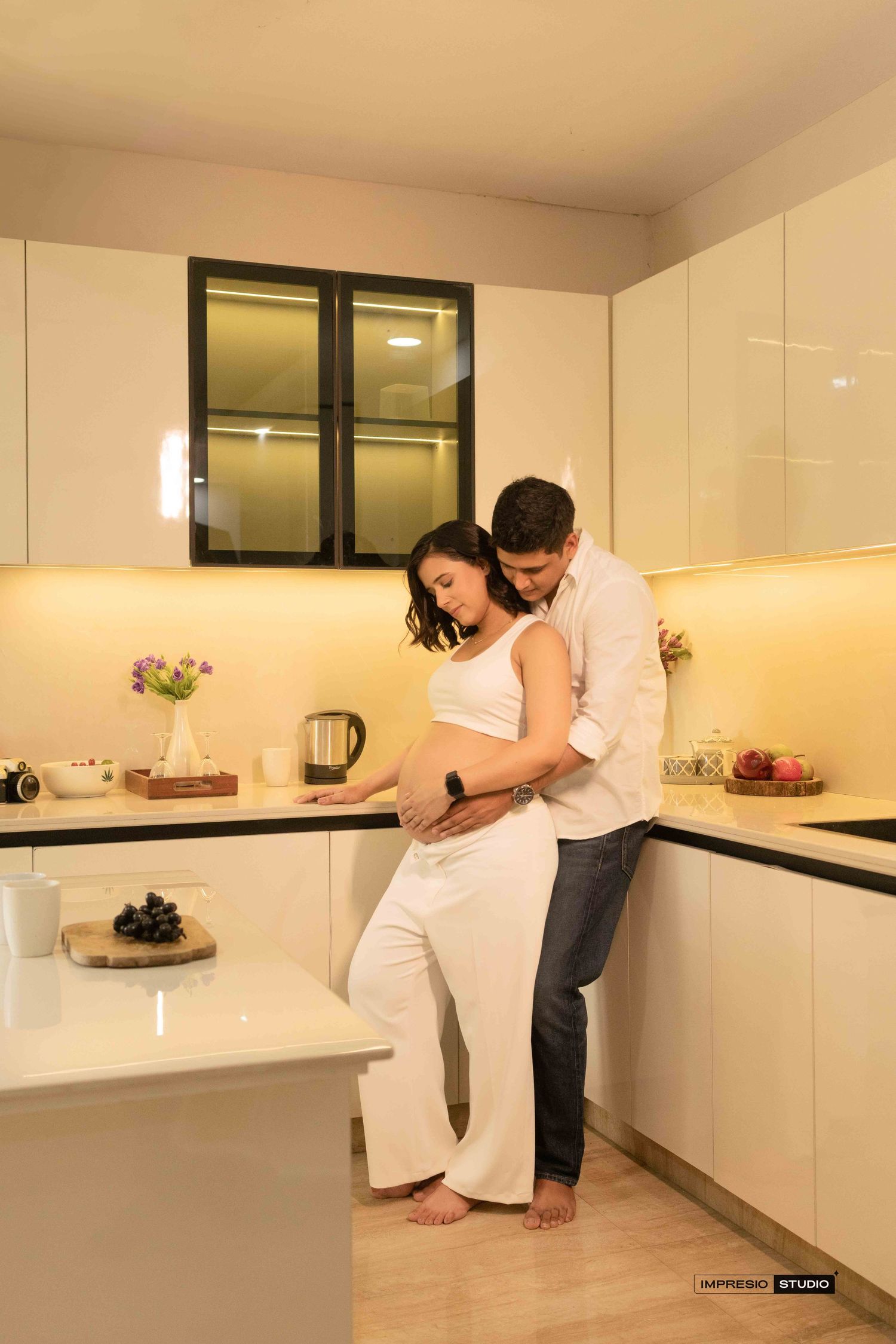 Couple sharing a tender maternity moment in a modern white kitchen, man embracing pregnant woman from behind.