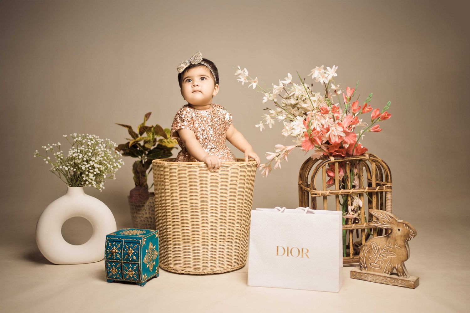 Baby girl in floral outfit sitting in wicker basket surrounded by flowers and a Dior bag in a styled photo shoot.
