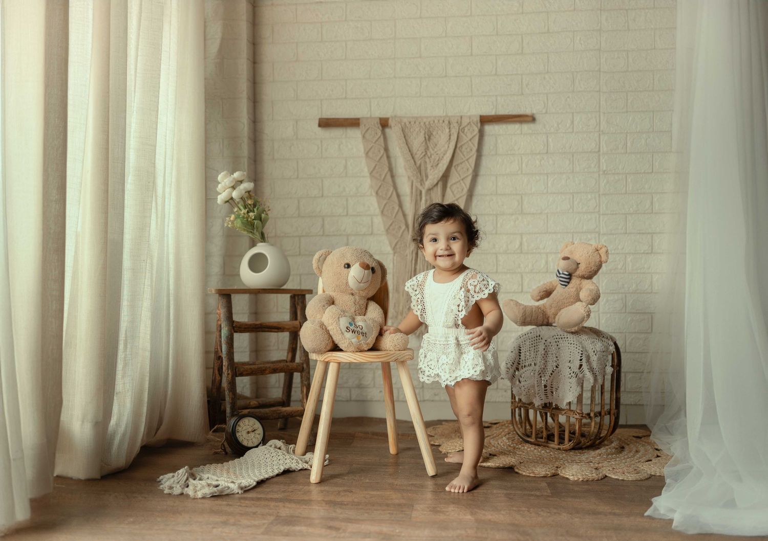 Toddler in white romper standing between two teddy bears in a bright, cozy nursery photo setup.