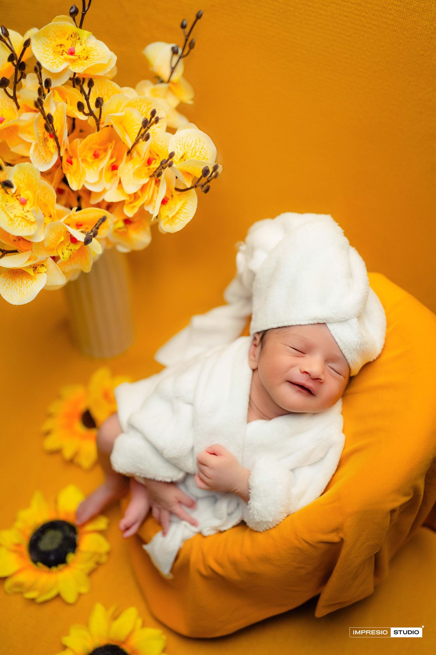 A smiling newborn baby sleeps in a white outfit surrounded by yellow sunflowers and orchids.