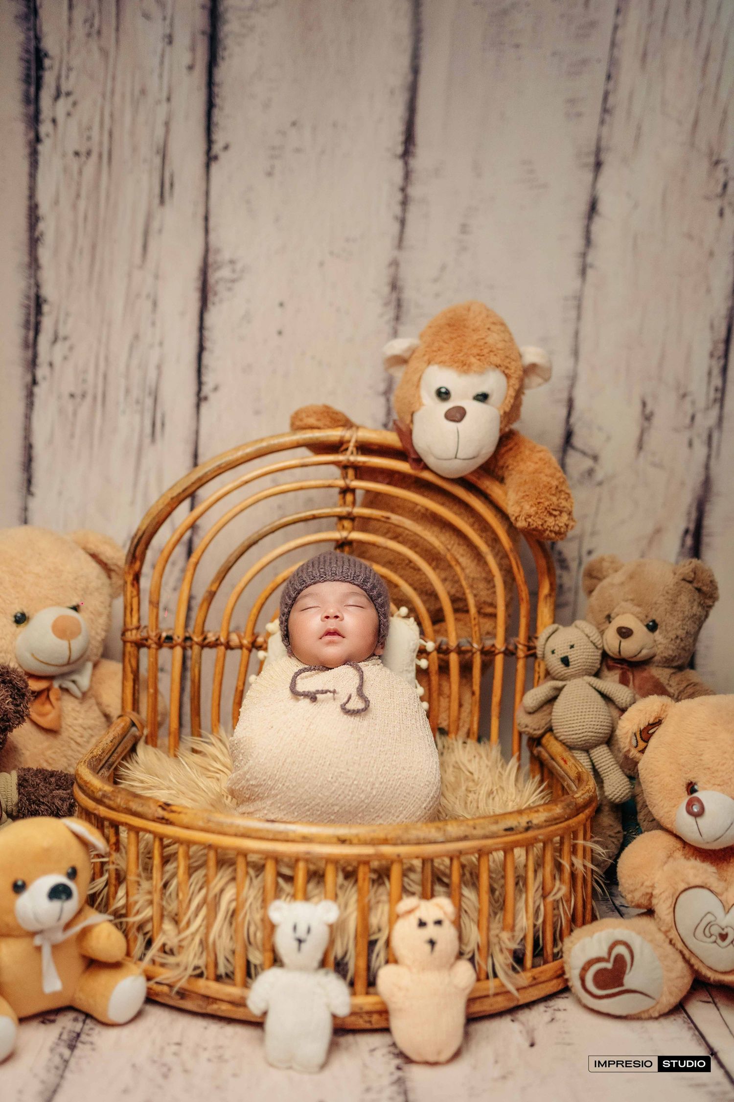 Newborn baby wrapped in white swaddle sleeping in wicker basket surrounded by teddy bears and stuffed animals.