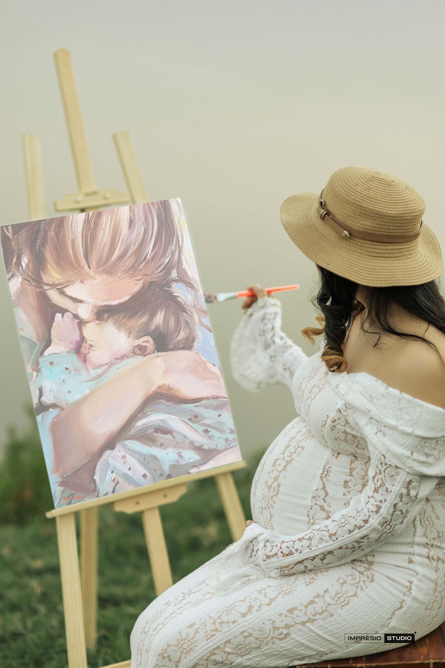 Pregnant woman in white lace dress and straw hat painting a mother-child portrait on an easel outdoors.